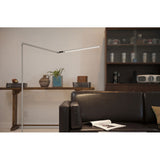 Silver Z-bar LED floor lamp lighting black leather sofa with newspaper resting on cushion. 